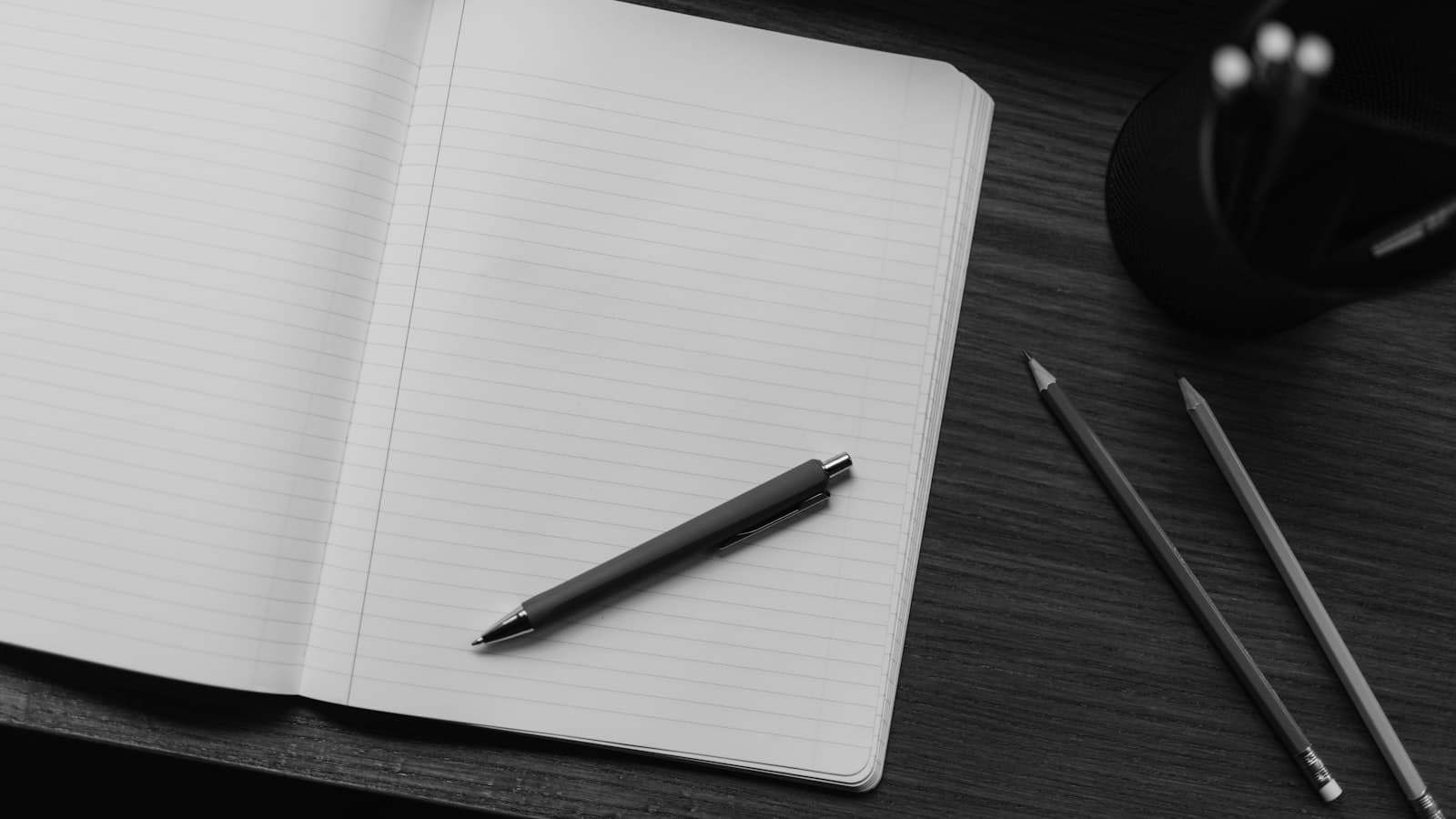 Black and white photograph of an open notebook with a pen and pencils on a desk